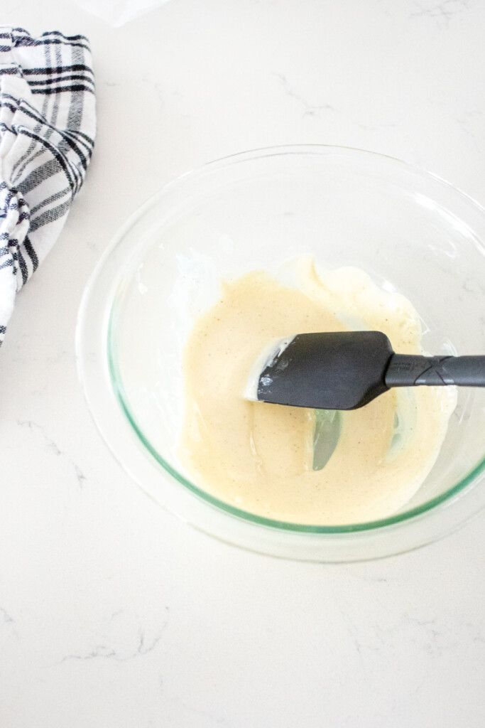 melted white chocolate in a glass mixing bowl with a spatula on a white marble counter.