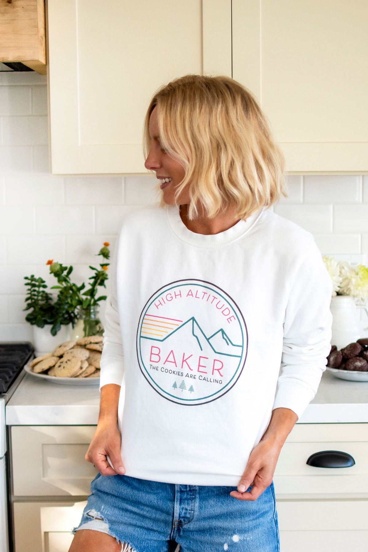 woman wearing a high altitude baking sweatshirt in the kitchen with cookie plates on the counter.