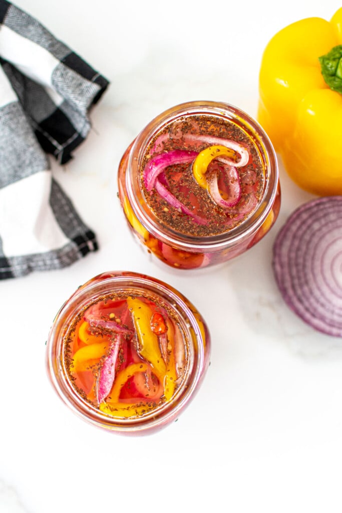 pickled onions and peppers in two mason jars on a white marble countertop.