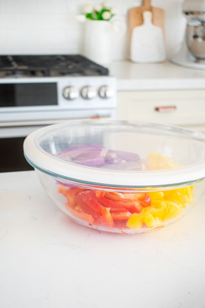 sliced red onion and bell peppers in a glass mixing bowl with a lid sitting on kitchen counter.