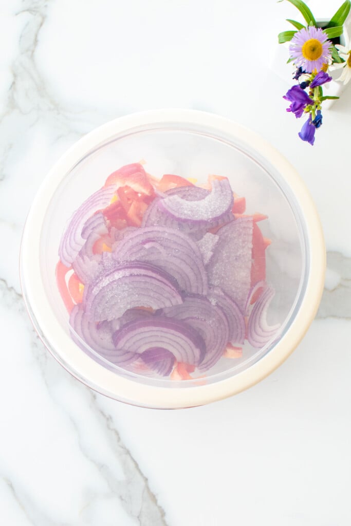 sliced red onion and bell peppers in a glass mixing bowl with a lid sitting on marble kitchen counter.