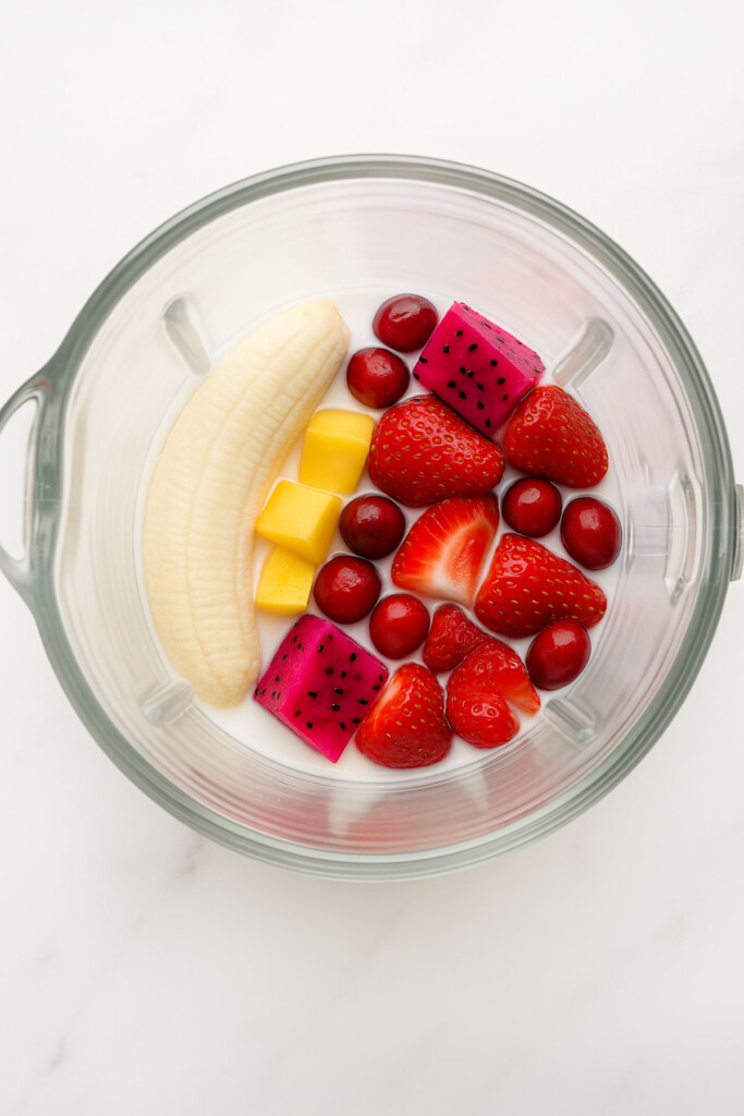ingredients for a tropical cranberry smoothie in a glass blender on a white marble counter.