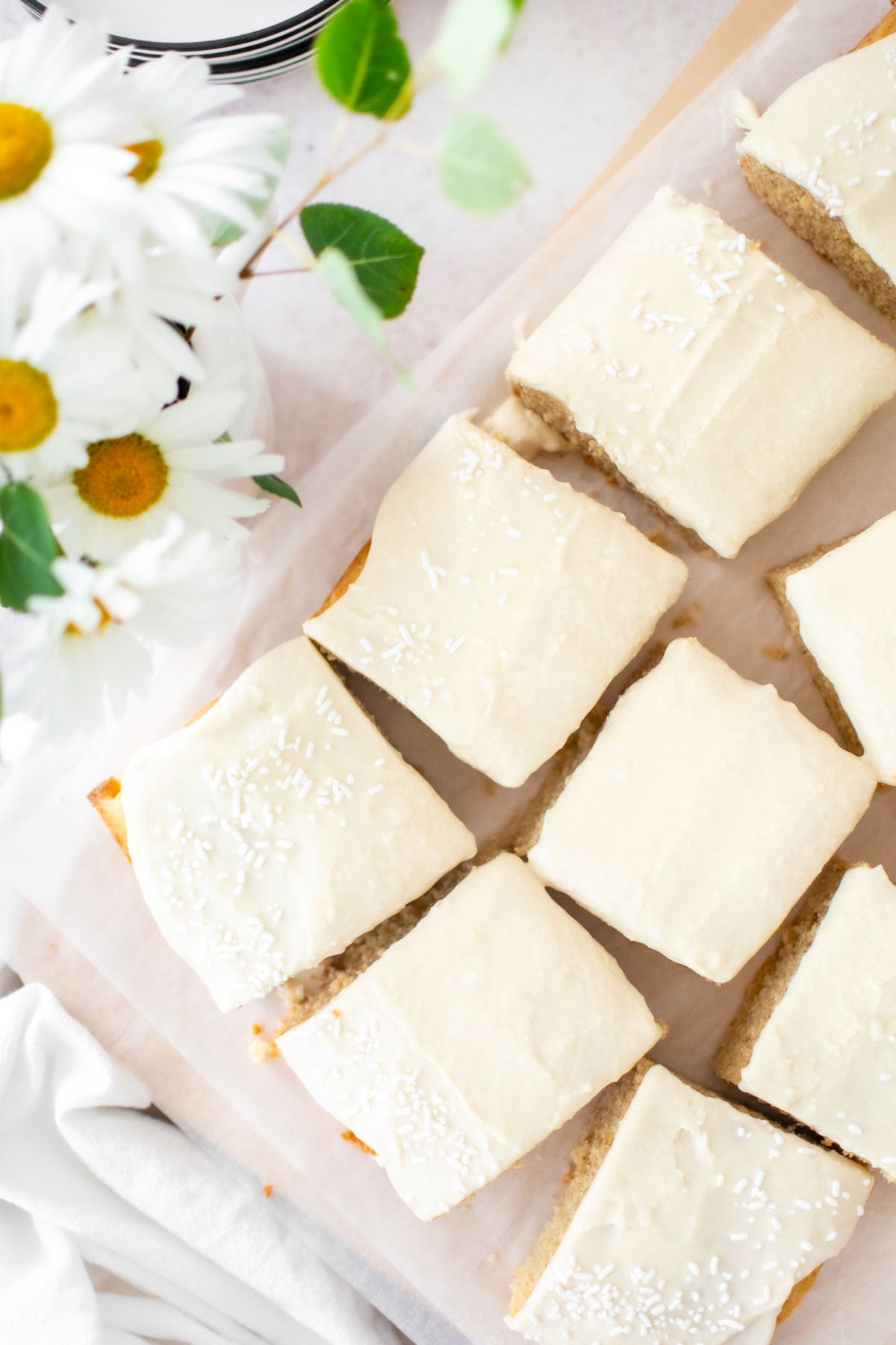 vanilla buttermilk sheet cake cut into square slices on a wood cutting board with fresh flowers on the table next to it.