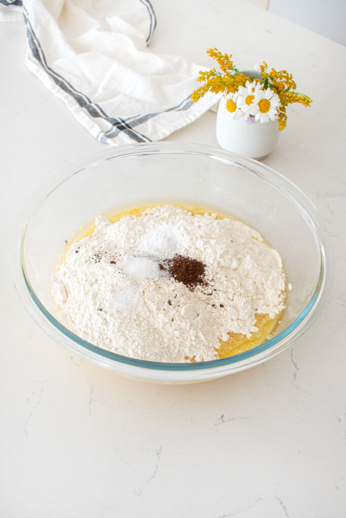 dry ingredients added to wet ingredients for vanilla buttermilk sheet cake in a glass mixing bowl on a marble counter with a vase of fresh flowers.
