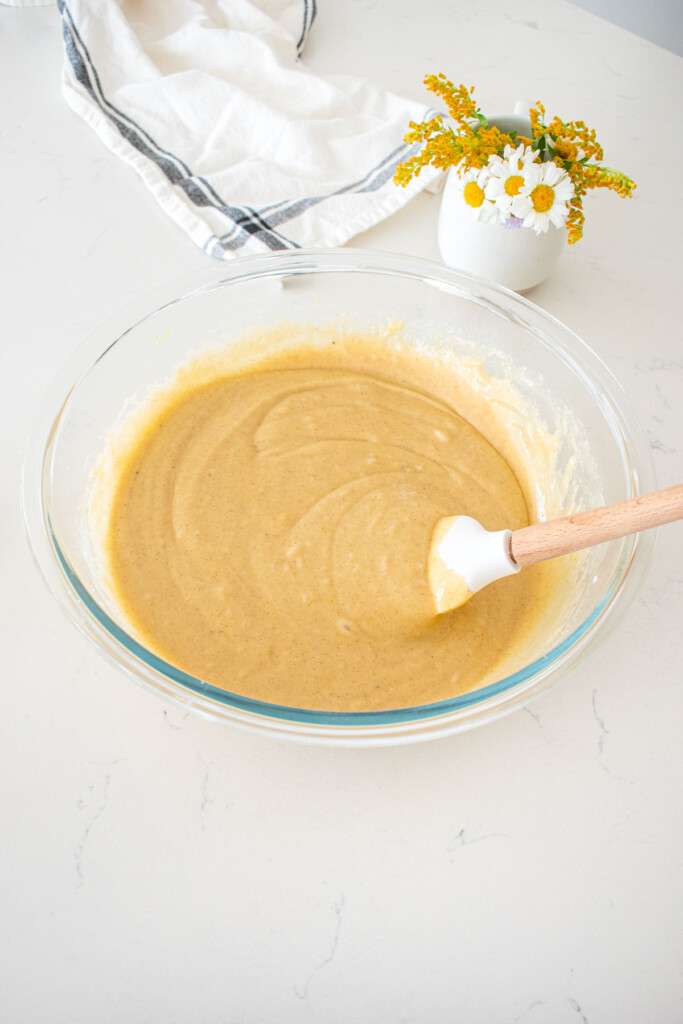vanilla buttermilk sheet cake batter in glass mixing bowl on a marble counter with a small vase of fresh flowers.