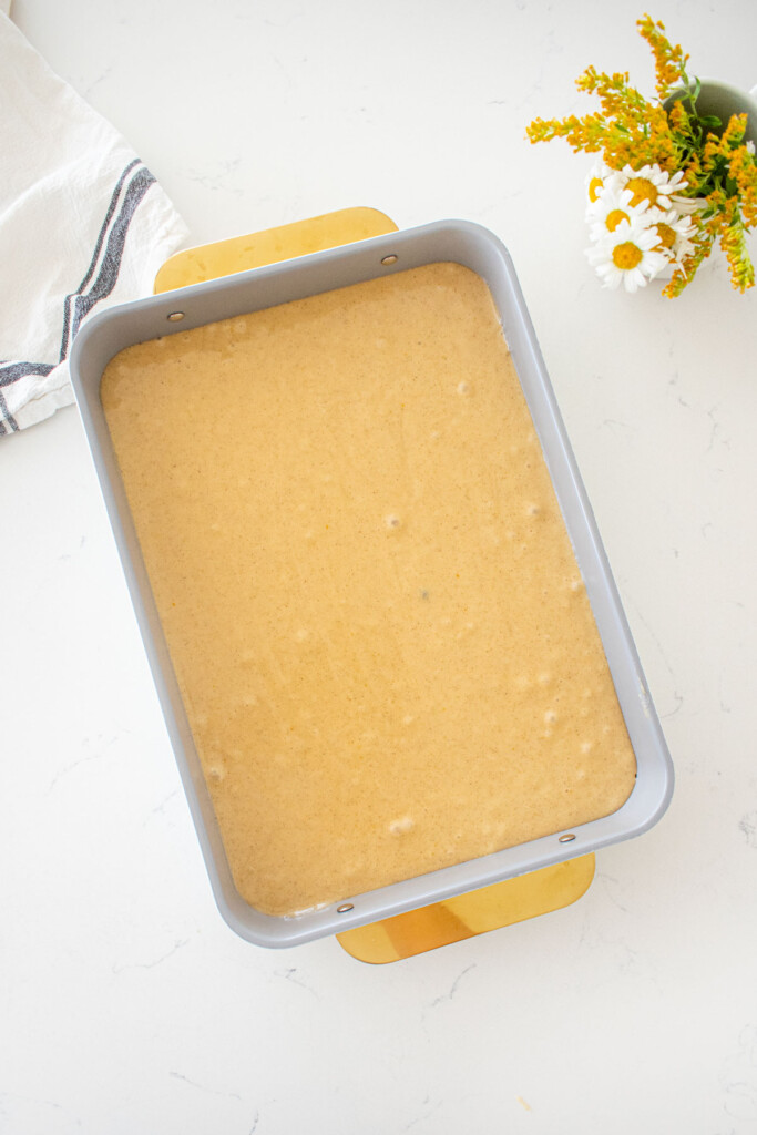 9x13-inch baking pan with vanilla buttermilk sheet cake batter spread evenly sitting on a marble counter with a vase of fresh flowers.