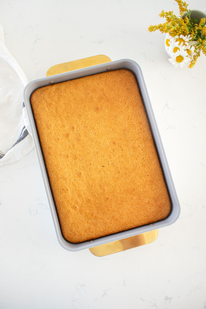 golden brown baked vanilla buttermilk sheet cake in a rectangle pan with gold handles on a marble counter with a vase of fresh flowers.