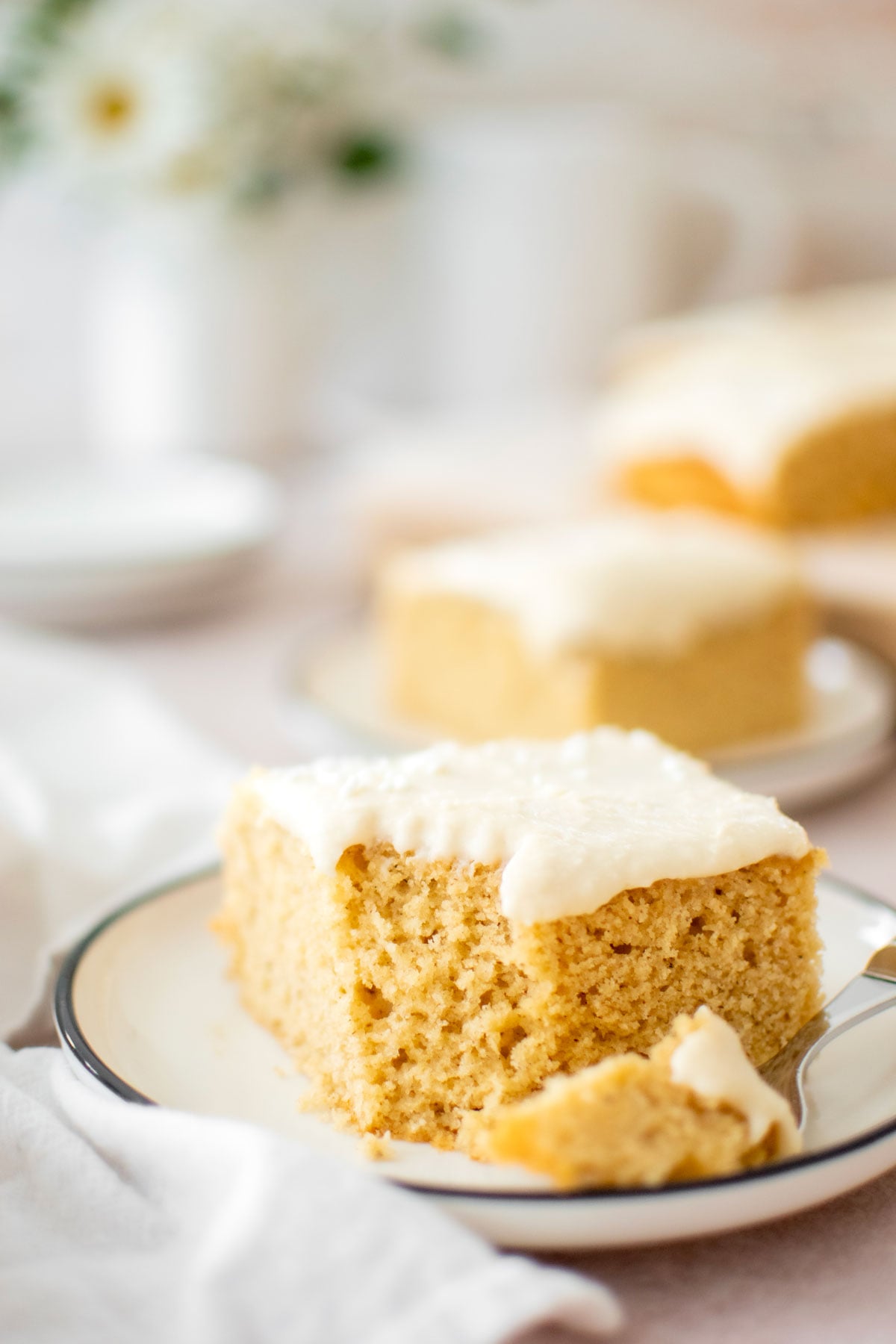 square slice of vanilla buttermilk sheet cake with vanilla frosting on a cake plate with a bite taken out.