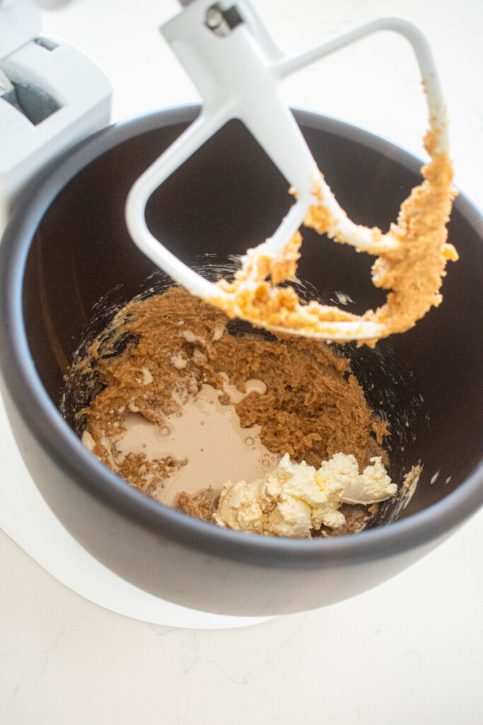 creamed butter, brown sugar and cane sugar in a wood mixing bowl with nondairy milk and cream cheese on a white marble counter.