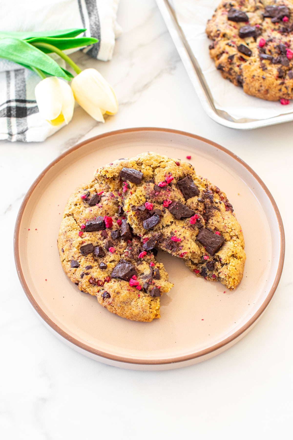 big vegan raspberry chocolate chip cookie broken in half on a plate on marble counter with flowers.