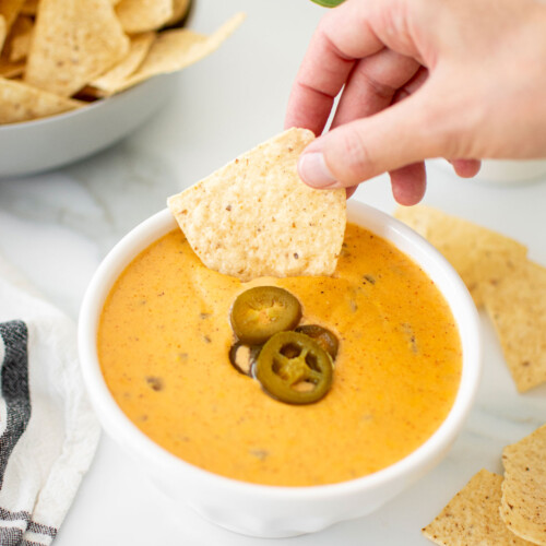bone broth queso dip in a white bowl, woman dipping a corn chip into it.