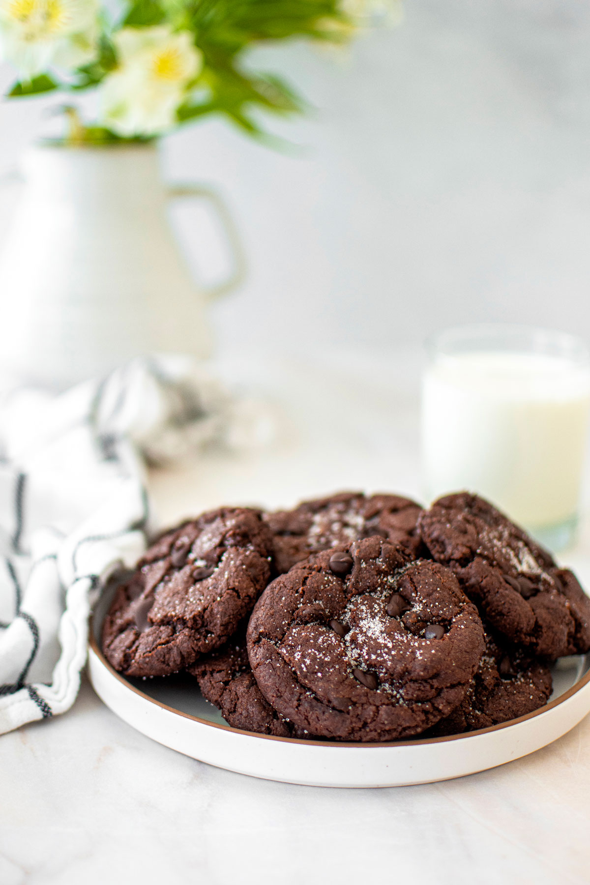 eggless chocolate chocolate chip cookies on a plate on a marble counter with a glass of milk and vase of flowers.