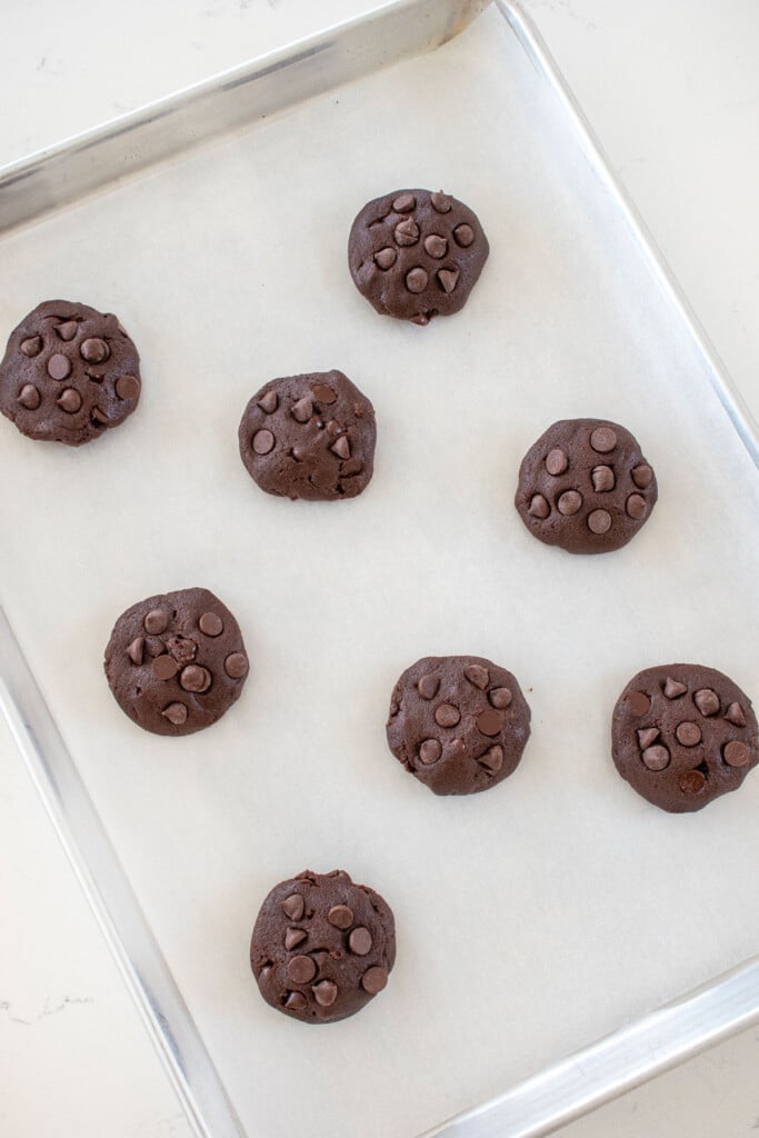 baking sheet with parchment paper and eggless chocolate chocolate chip cookie dough in balls on a marble countertop.