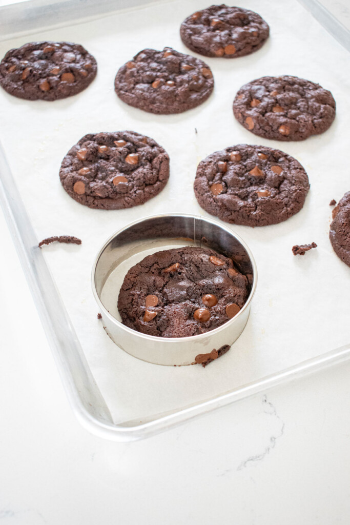 eggless chocolate chocolate chip cookies on a baking sheet being rounded out with a cookie cutter after baking.
