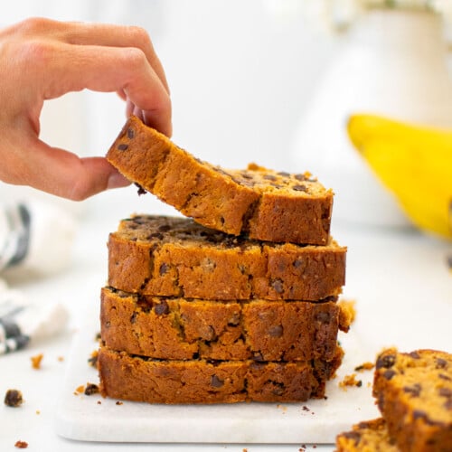eggless mini chocolate chip banana bread slices stacked on a white marble cutting board, woman grabbing the top slice.