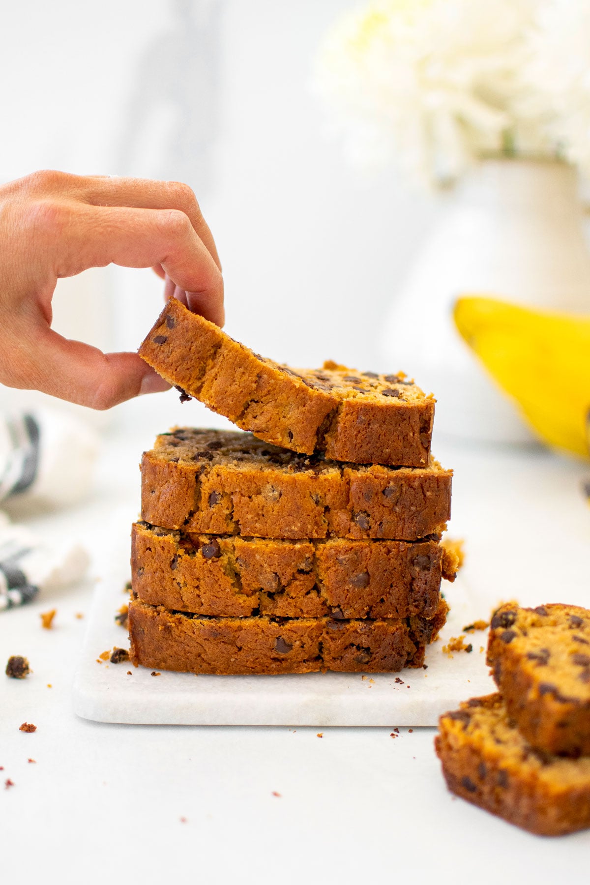 eggless mini chocolate chip banana bread slices stacked on a white marble cutting board, woman grabbing the top slice.