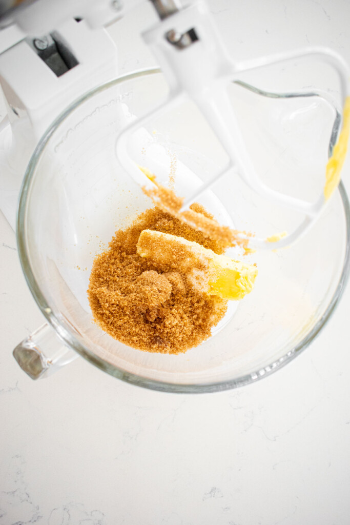 butter, sugar and brown sugar in a glass mixing bowl on a white marble countertop.
