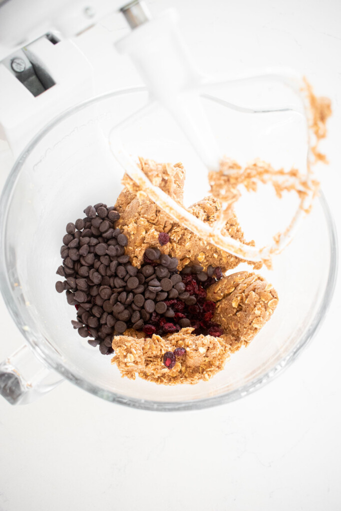 chocolate chips and cranberries being added into eggless oatmeal cookie dough in a glass mixing bowl on a marble countertop.