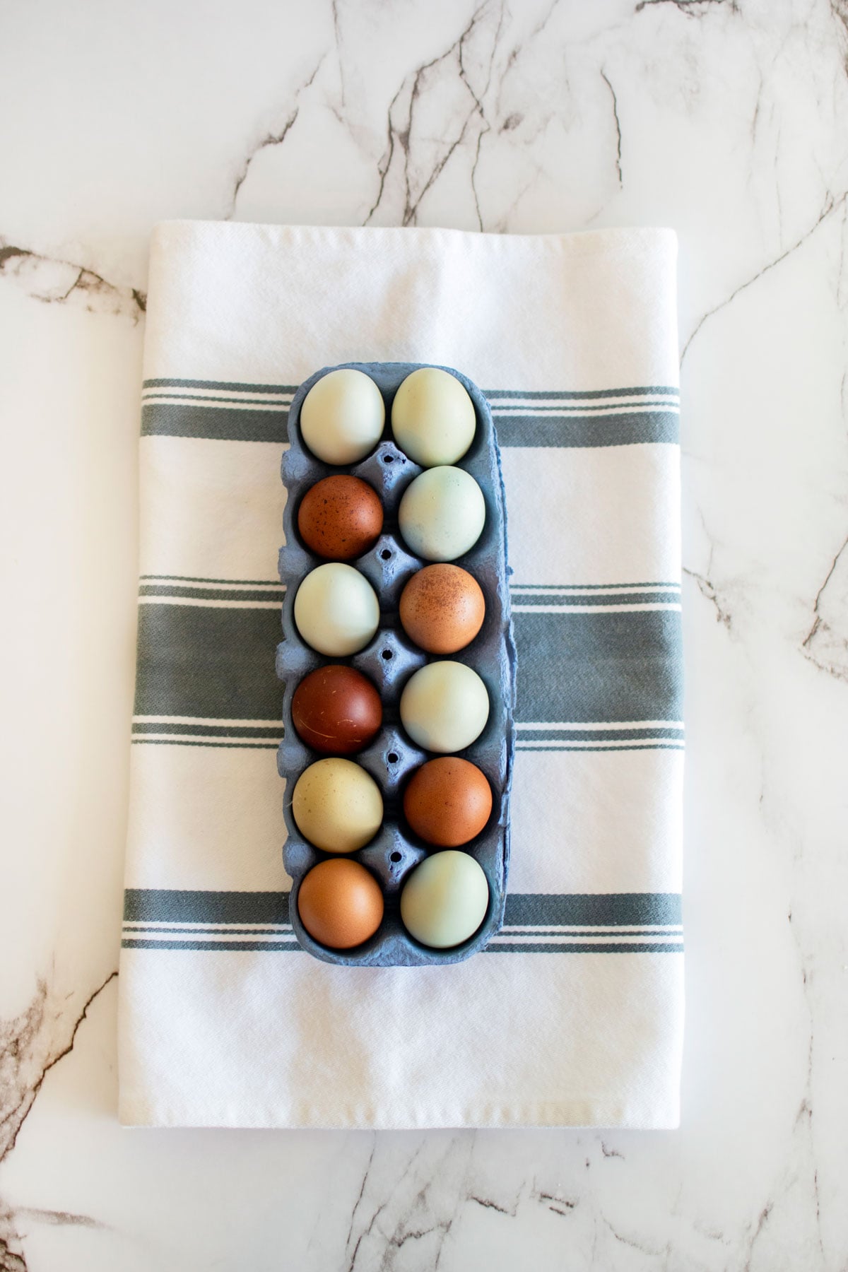 brown, green, and blue farm eggs in a blue egg carton on a towel on marble countertop.