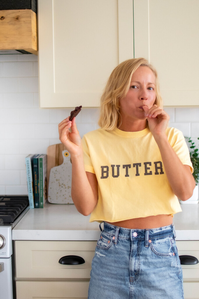 woman wearing a butter tee shirt eating an eggless chocolate chocolate chip cookie in the kitchen.