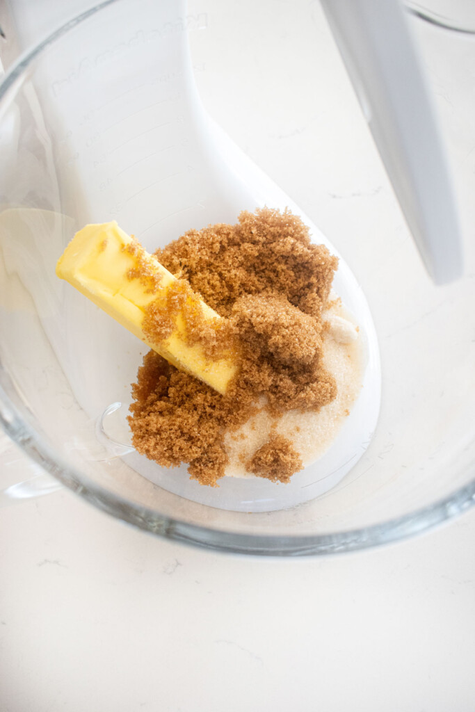 butter and brown sugar in a glass stand mixing bowl on a white marble counter.