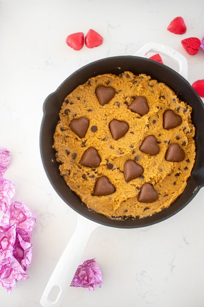 peanut butter cookie dough with chocolate hearts pressed into it in a skillet on a marble counter.