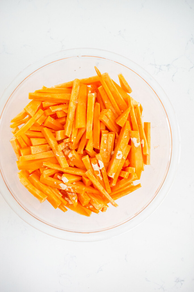 sweet potato cut into fries in a glass bowl with garlic powder and lemon on a marble countertop.