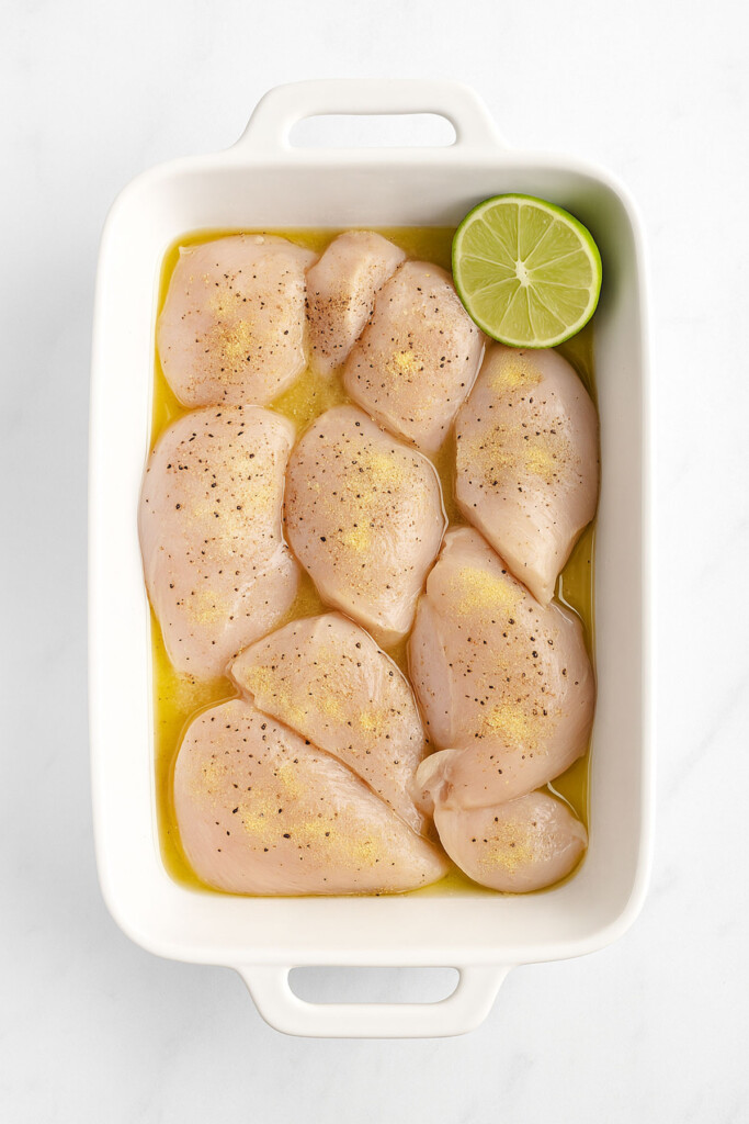raw chicken breasts marinating in a baking dish with spices and lime on a white marble countertop.