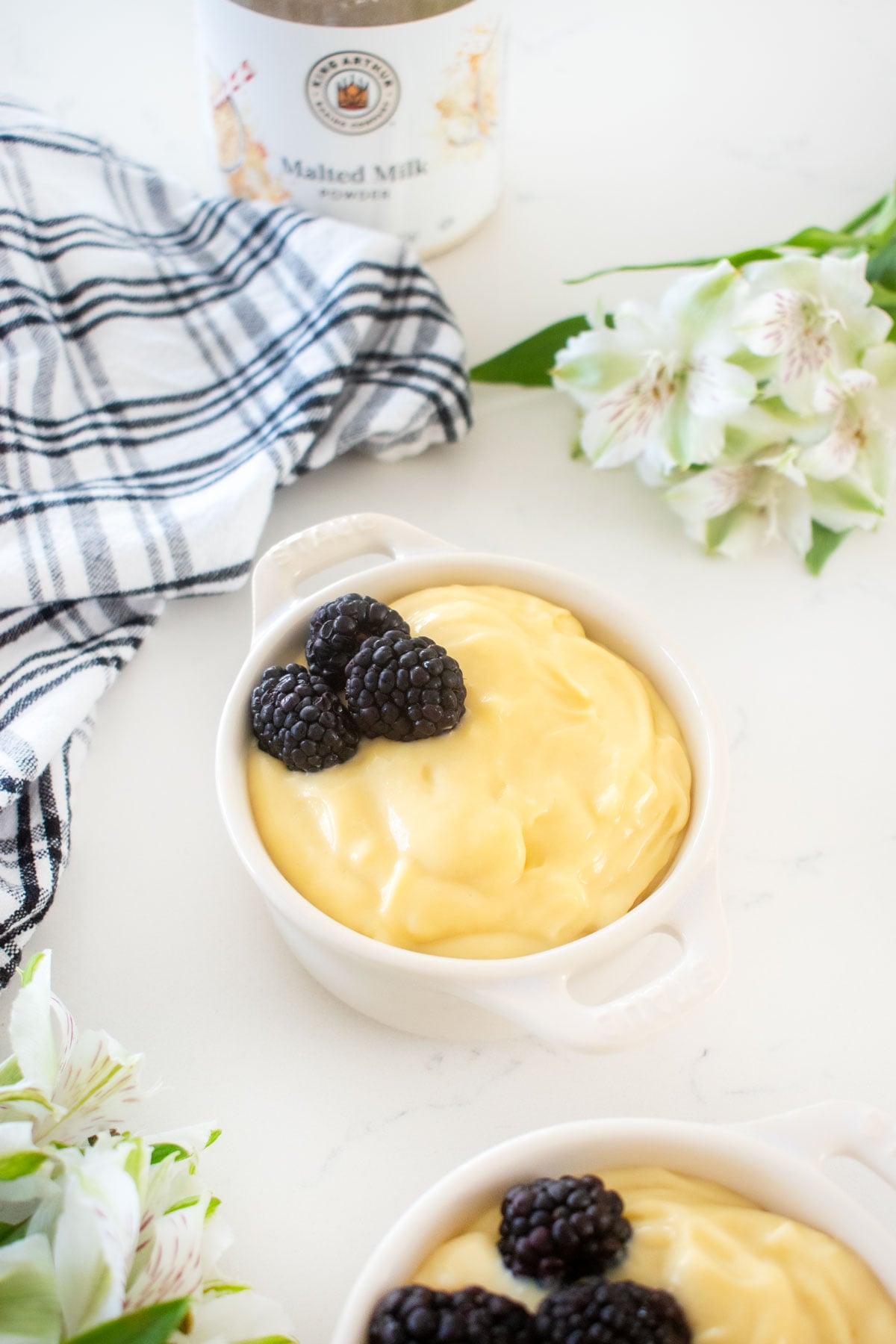 homemade vanilla malt pudding in a mini white dutch oven with fresh blackberries on a white marble counter with flowers.