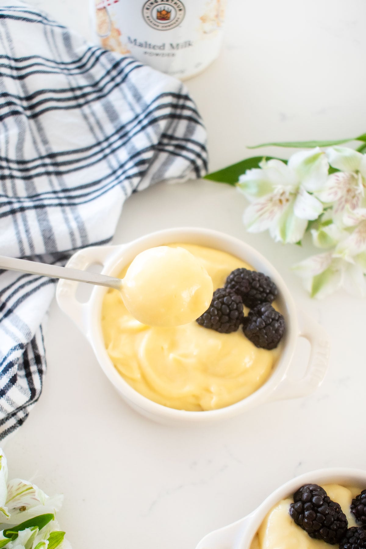 spoonful of homemade vanilla malt pudding coming out of a ramekin with fresh blackberries on a white marble counter with fresh flowers.