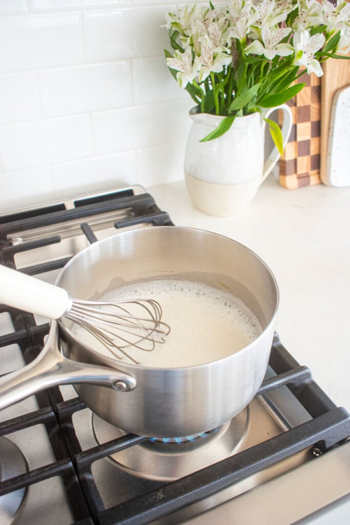 stainless steel pot of milk and sugar on a stovetop being whisked.