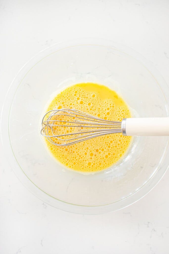 glass mixing bowl with egg yolks whisked with milk on a white marble counter.