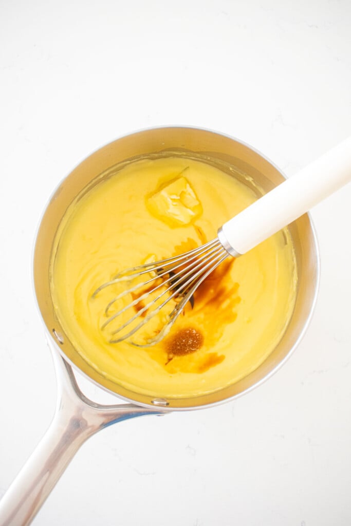 homemade vanilla malt pudding being whisked with butter and vanilla in a pot on a white marble counter.