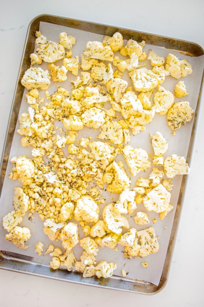 raw cauliflower pieces with seasoning spread out onto a baking sheet on a marble counter.