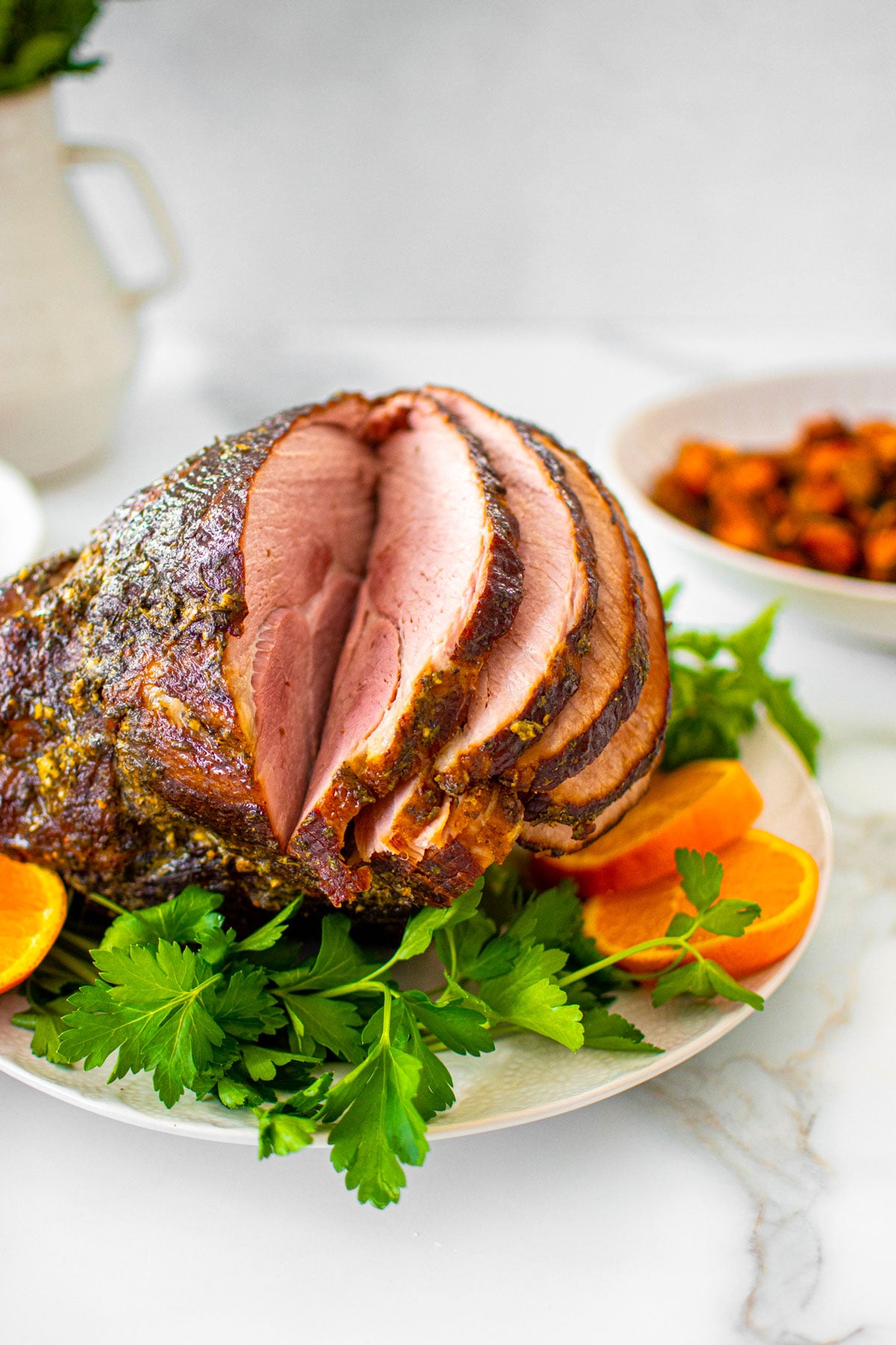 savory mustard herb ham on a platter with fresh parsley and orange slices on a white marble table with holiday sides.