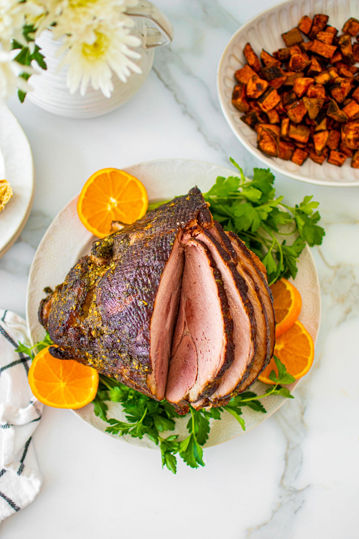 savory mustard herb ham on a platter with fresh parsley and orange slices on a white marble table with holiday sides.
