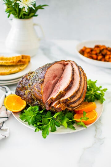 savory mustard herb ham on a platter with fresh parsley and orange slices on a white marble table with holiday sides.