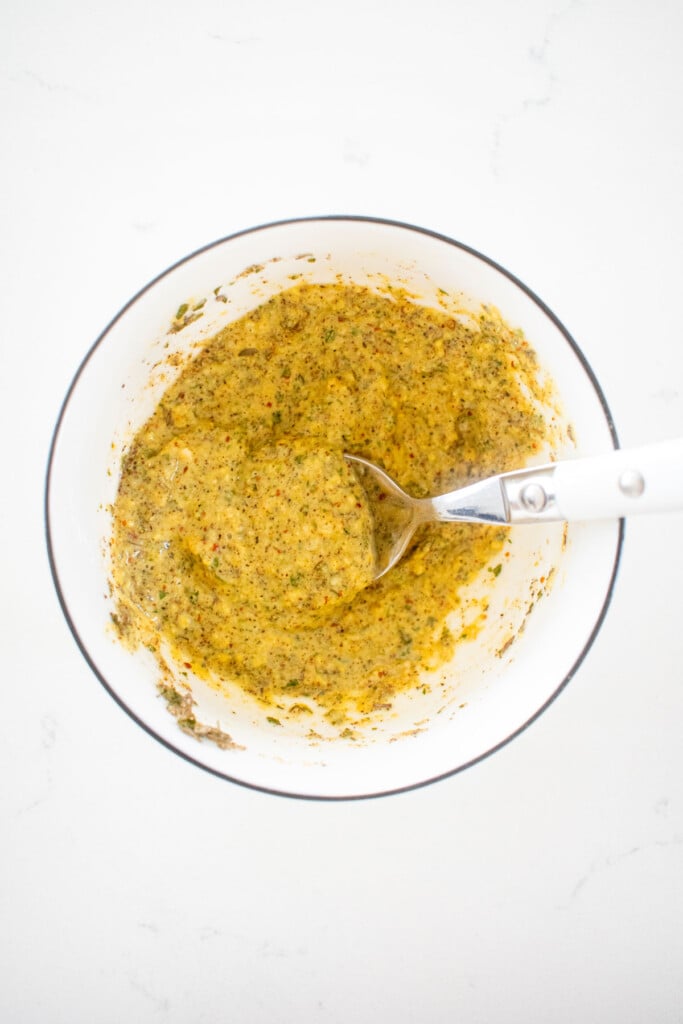 herbs and spicy brown mustard mixed together in a small bowl on a white marble counter.