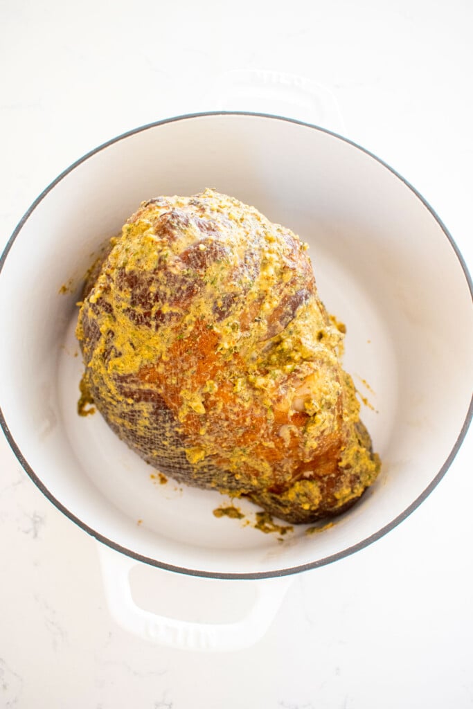 savory mustard herb ham in a white dutch oven with spice rub all over it on a marble counter.
