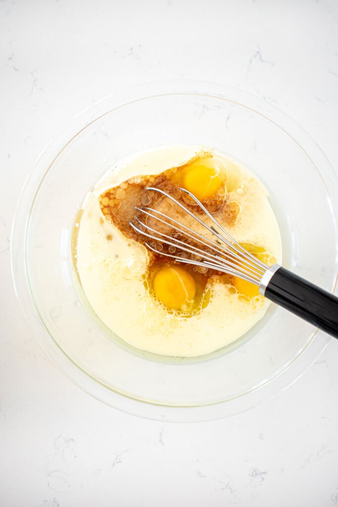 ingredients for vanilla cake batter in a glass mixing bowl with a whisk on a white marble counter.
