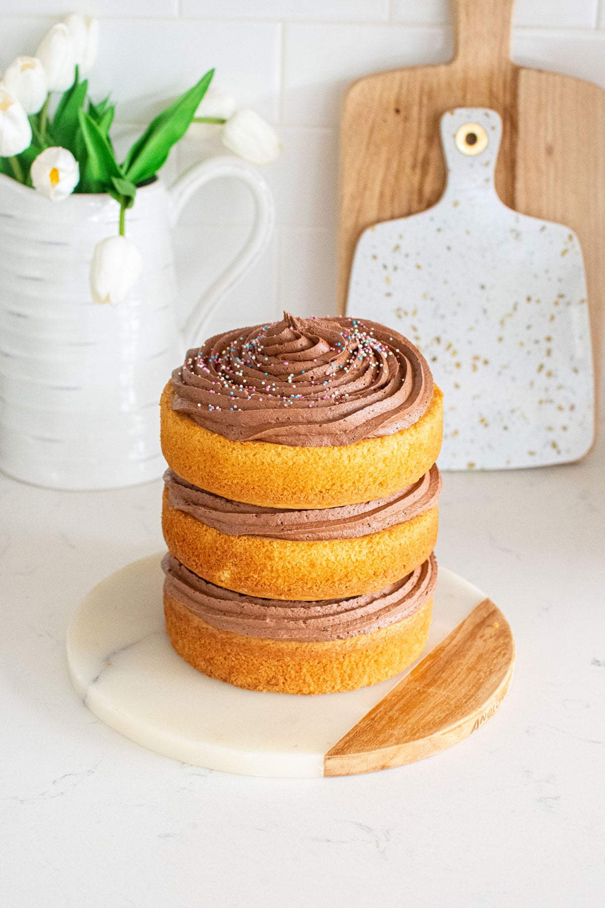 simple naked birthday cake with chocolate frosting and rainbow sprinkles on a marble cake plate on a white kitchen counter.