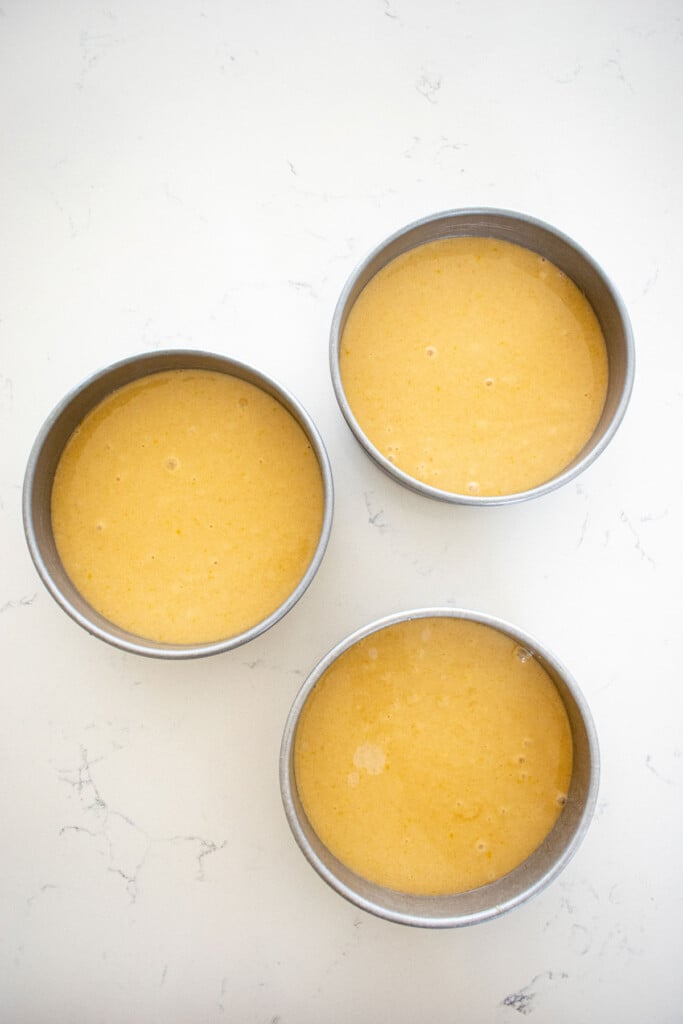 vanilla cake batter in three 6-inch cake pans on a white marble counter.