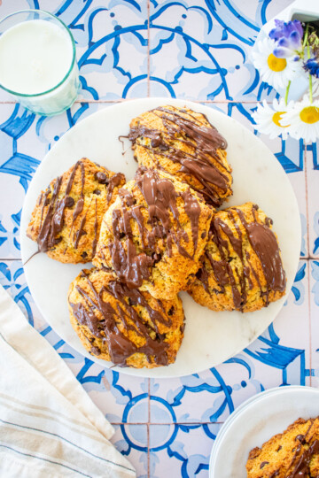marble serving board with sourdough chocolate chip scones with melted chocolate drizzle on top on a tile table with a glass of milk and fresh flowers.