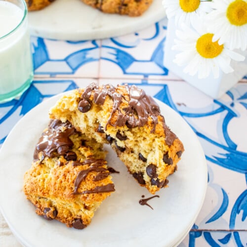 sourdough chocolate chip scone with melted chocolate drizzle on top broke in half on a plate on a tile table with flowers.
