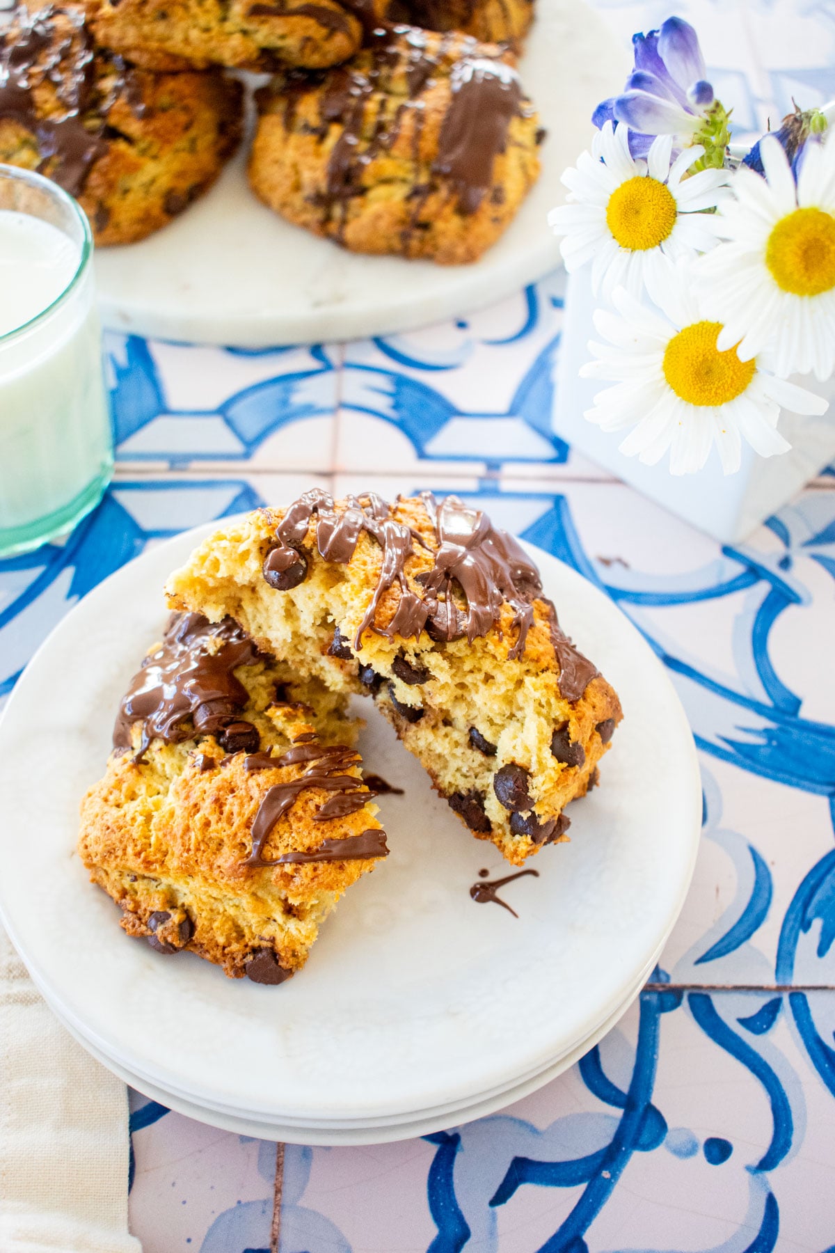 sourdough chocolate chip scone with melted chocolate drizzle on top broke in half on a plate on a tile table with flowers.