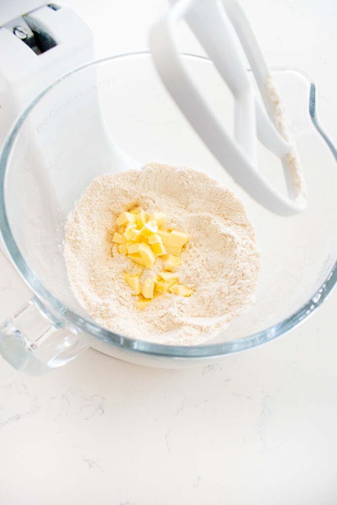 chopped butter added into flour in a glass stand mixing bowl on a white marble counter.