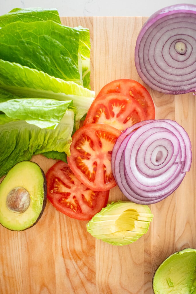 wood cutting board with lettuce leaves, sliced tomato, sliced red onion and sliced avocado.
