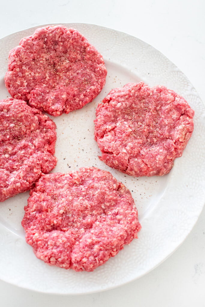 plate with four burger patties formed with salt and pepper on a white marble counter.