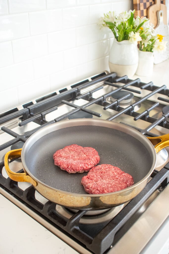 large skillet with two burger patties being cooked on the stovetop.