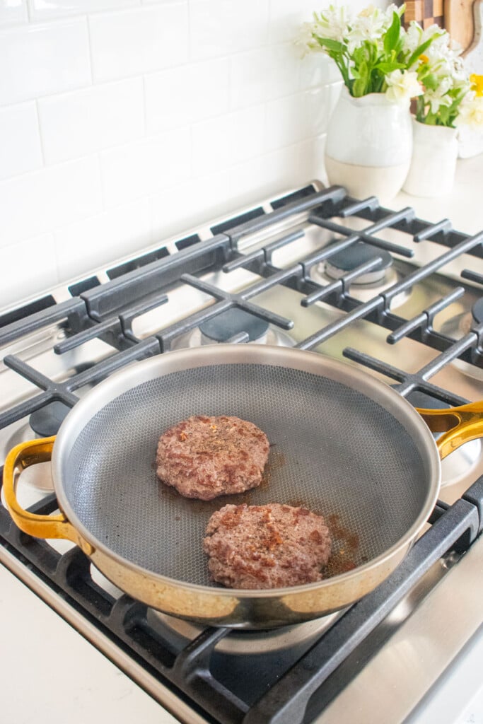 large skillet with two burger patties being cooked on the stovetop.
